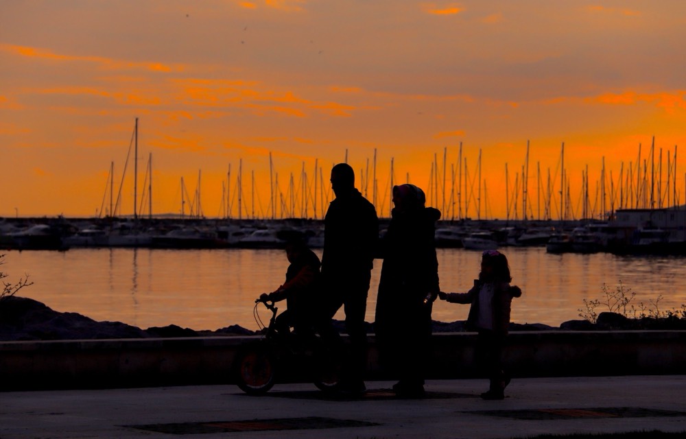 A family walking together along a harbor at sunset — one parent steadying a child’s bike, another holding their daughter’s hand